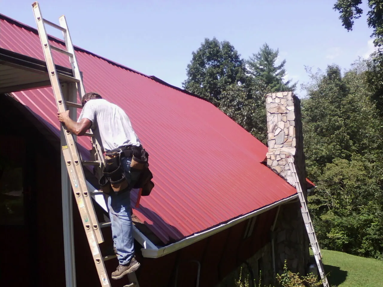 Roofer on a ladder beside a red metal roof and stone chimney — Soffit & Fascia Repair by Roofing Brothers in Lewisburg